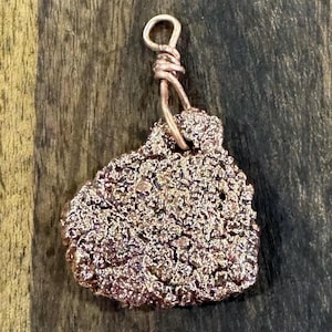 May include: A copper-colored pendant with a textured, irregular surface. The pendant is suspended from a looped wire bail. The pendant's surface has a rough, porous appearance, and the overall design is organic and earthy.