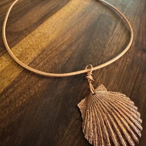 Puede incluir: Un collar de color cobre con un colgante de concha. El colgante tiene líneas texturizadas detalladas y cuelga de un choker redondo y simple. El collar se presenta sobre una superficie de madera.