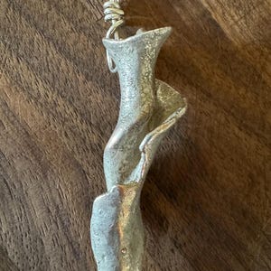 May include: A silver-colored pendant with a unique, abstract design. The pendant has a flowing, organic shape with textured surfaces and a loop at the top for hanging. The piece is displayed against a dark wood grain background.