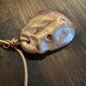 Pode incluir: Um pingente de bronze metálico com uma forma natural e orgânica. O pingente tem vários pequenos orifícios e é preso a um fio de bronze fino e curvo. O fio é conectado a um elemento de bronze mais espesso e enrolado.