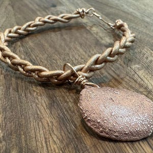 May include: A copper-colored necklace with a braided chain and a teardrop-shaped pendant. The pendant has a textured surface. The necklace is displayed on a wooden surface.