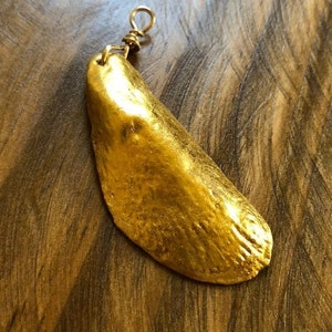 Pode incluir: Um pingente dourado com um design texturizado em forma de folha. O pingente é suspenso por um pequeno laço de arame dourado enrolado. O fundo é uma superfície de madeira com grão visível.