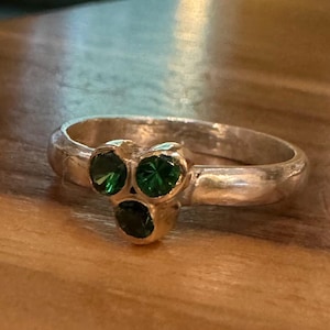 Könnte beinhalten: Ein silberner Ring mit drei smaragdgrünen Edelsteinen. Der Ring hat ein abgerundetes Band, und die Edelsteine sind in einer Gruppe angeordnet. Der Ring wird auf einer Holzoberfläche gezeigt.