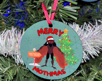 May include: A round, green Christmas ornament with a white snowflake pattern. It features a cartoon moth wearing a Santa hat, standing in front of a Christmas tree with the text "Merry Mothmas" and a sign that reads "Point Pleasant, West Virginia".
