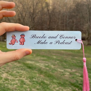 May include: White bookmark with a pink tassel. The bookmark features two illustrated teddy bears wearing a crown and a baseball cap. The text "Brooke and Connor Make a Podcast" is written in cursive.