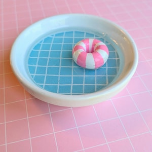 May include: A round white ceramic dish featuring a blue and white grid pattern on the interior base, resembling a swimming pool. A small, circular, pink and white striped inflatable ring float sits in the center of the dish, resting on the base.