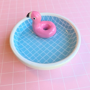 May include: A round ceramic trinket dish featuring a light blue interior with a white grid pattern resembling a swimming pool. A small, pink, flamingo-shaped pool float is attached to the center of the dish, complete with a black and white beak.