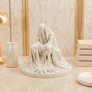 Peut inclure: Une sculpture blanc mat représentant une silhouette assise drapée dans un tissu fluide qui recouvre la tête et le corps, reposant sur une base circulaire. La silhouette présente des bras et des jambes musclés sous les plis du tissu texturé.