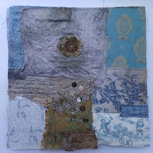 May include: A square textile collage featuring various fabric patches in shades of blue, grey, and mustard yellow. The piece includes a central metallic circular element, intricate lace trim, and several small buttons sewn onto the fabric surface.