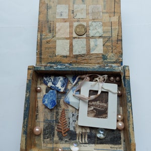 May include: An open wooden box with a blue interior. The box is filled with various items, including small pieces of blue and white china, a fern, a small photo, and a few white beads. The top of the box is decorated with a grid of small squares, some of which are filled with old postage stamps.