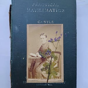 May include: A dark green hardback book titled Practical Mathematics by Castle, featuring a vintage sepia-toned photograph of a person seated at a table, overlaid with a pressed purple flower stem and green leaves on the front cover.