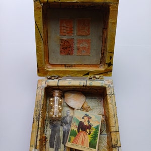 May include: A small wooden box with a hinged lid, decorated with paper scraps, stamps, and a seashell. Inside the box, there is a small glass bottle filled with white beads, a postcard with a woman in traditional Austrian dress, and a man in a suit.