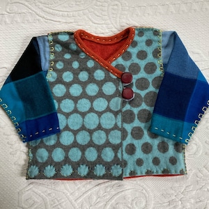 Puede incluir: Una chaqueta cruzada de manga larga con un patrón de lunares azul claro y gris en la parte delantera. Las mangas tienen un diseño de cuadros azules y negros con costuras decorativas amarillas. Incluye dos botones rojo oscuro y forro naranja.