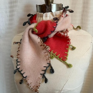 May include: A handmade, colorful scarf displayed on a mannequin. The scarf features a patchwork design with shades of pink and red, accented with decorative stitching and small, dangling embellishments. The scarf is made of a soft, knitted material.