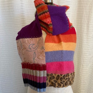 May include: A colorful patchwork scarf displayed on a mannequin. The scarf features various textures and patterns, including stripes, floral embroidery, and a leopard print section. The color palette includes shades of purple, orange, pink, and brown.