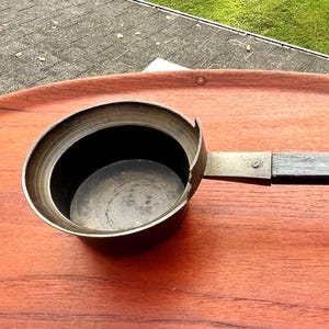 May include: A metal ladle with a deep, circular bowl and a long, straight, dark-colored handle. The bowl features a distinct notched rim on one side, and the handle is attached to the bowl with a visible metal fastener, showing a weathered finish.