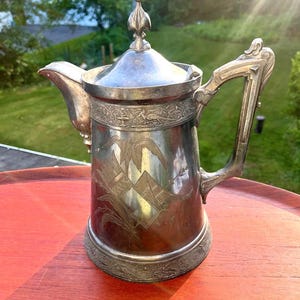 May include: A silver-toned metal coffee pot featuring a tapered cylindrical body, a hinged lid with a pointed finial, and an ornate handle. The exterior surface is decorated with etched botanical motifs and a band of intricate relief patterns.