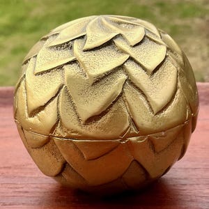 May include: A golden-colored, spherical decorative box with a textured, overlapping leaf design. The box is made of metal and has a hinged opening. The box is sitting on a wooden surface.