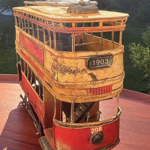 May include: A metal model of a double-decker tram painted in red and yellow with a weathered finish. The front displays the number 208 and the upper level features a black sign with the year 1903. It includes open windows and a roof structure.