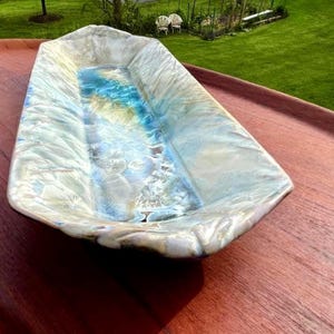 May include: An elongated, rectangular ceramic tray featuring a crystalline glaze with a glossy finish. The surface displays a blend of iridescent white, cream, and soft blue tones, with intricate, star-like patterns radiating across the interior.