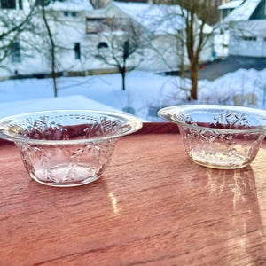 May include: Two clear glass bowls with a decorative floral pattern are displayed on a reddish-brown wooden surface. The bowls have a flared rim and a detailed design. The background shows a snowy landscape with houses and trees.