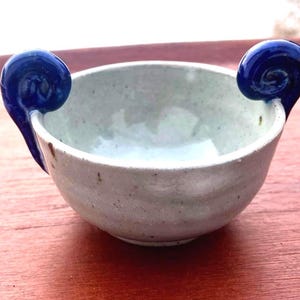 May include: A small, round ceramic bowl with a speckled off-white glaze and two prominent, glossy cobalt blue handles shaped like curled ram horns attached to opposite sides of the rim. The bowl has a smooth, slightly uneven texture.
