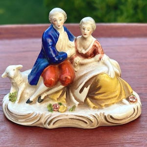 May include: A porcelain figurine depicting two figures seated together with a small lamb. The figure on the left wears a blue coat and orange breeches, while the figure on the right wears a red bodice and a yellow skirt, all on a base.