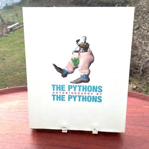 May include: A white book cover titled THE PYTHONS AUTOBIOGRAPHY BY THE PYTHONS in blue and red text. The center graphic depicts a person's upper body emerging from a pair of large, walking legs wearing brown shoes, blue socks, and suspenders.