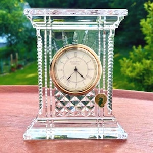 May include: A clear crystal mantel clock with a rectangular frame featuring vertical fluted edges and a diamond-cut base. The circular clock face has a gold-tone bezel, black hands, and Roman numerals with the text Waterford Crystal printed on it.