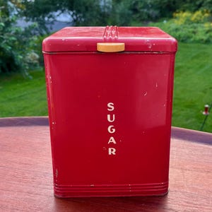May include: A rectangular red metal canister with a hinged lid and a small yellow tab handle. The word SUGAR is printed vertically in white capital letters on the front face. The container features horizontal ribbed detailing at the base.