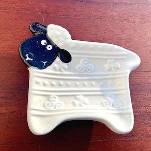 May include: A white ceramic sheep-shaped dish with a black and white head. The dish has a curved shape with embossed details, including horizontal lines and floral designs. The sheep's face has two white eyes and a small, white mouth.