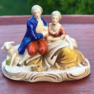 May include: A porcelain figurine depicting a couple in 18th-century attire, seated on a cloud-shaped base. The man wears a blue coat and red breeches, while the woman is in a red and yellow dress. A white lamb rests beside them. The figurine is painted in soft, muted colors.