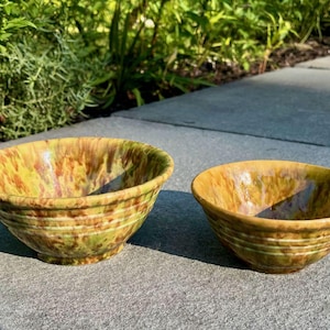 May include: Two ceramic bowls with a speckled glaze in shades of yellow, green, and brown. The bowls have a rounded shape with a slightly flared rim and horizontal bands around the exterior. They are placed on a gray surface.