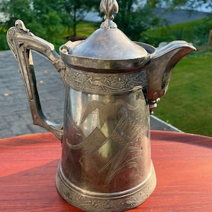 May include: Silver-plated antique coffee pot with a lid and ornate handle. The pot features engraved designs of birds, wheat, and geometric patterns. The pot is approximately 10 inches tall.