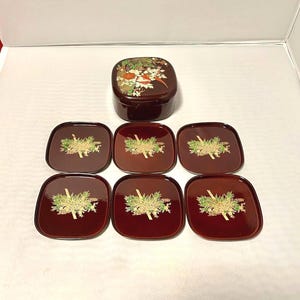 May include: A set of Japanese lacquerware. The set includes a dark brown, square-shaped box with a floral design on the lid, and six matching square coasters. Each coaster features a gold and green grass design.