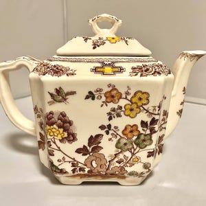 May include: Cream-colored ceramic teapot with a lid, handle, and spout. The teapot is decorated with brown floral and bird patterns, with accents of yellow, orange, and green. The teapot has a square shape and a small handle on the lid.