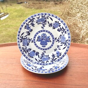 May include: Two white ceramic plates with a blue floral design. The plates feature a central basket motif surrounded by flowers and leaves. The rims are decorated with a geometric pattern. The plates are on a wooden surface.