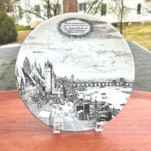 May include: A white decorative plate with a black and white illustration of a cityscape, including buildings, a bridge, and boats. The plate has text at the top that reads "FRANCOFURTUM UT". The plate is displayed on a clear stand.