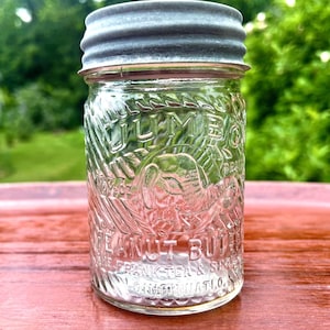 May include: A clear glass jar with a grey metal lid. The jar features embossed text reading JUMBO BRAND PEANUT BUTTER, THE FRANK TEA & SPICE CO. CINCINNATI O., and 10 OZ. A raised elephant graphic is centered on the front of the glass container.