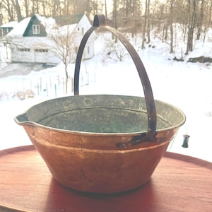 May include: A copper-colored metal cauldron with a curved handle. The cauldron has a wide, rounded bowl shape and a slightly flared rim. The handle is made of a darker metal and is attached to the sides of the cauldron. The background shows a snowy landscape.