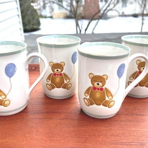 May include: Four white ceramic mugs with a light blue rim and handle. Each mug features a brown teddy bear with a red bow tie and a blue balloon. The mugs are on a wooden surface.