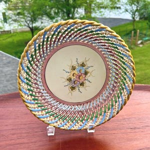 May include: A decorative plate with a woven, basket-like design. The rim features a mix of colors including yellow, blue, green, and red. The center of the plate has a floral design with pink, blue, and yellow flowers.