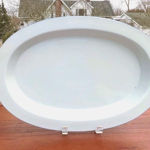 May include: A large, plain, oval-shaped white serving platter with a smooth, glossy finish and a wide rim. The platter is positioned upright on two small, clear acrylic stands, showcasing its clean, minimalist design and bright white color.