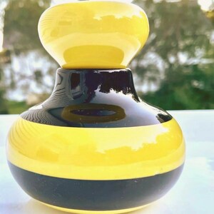 May include: A small, rounded ceramic vessel featuring a glossy finish with alternating horizontal bands of bright yellow and deep black. The object has a wide, bulbous base that tapers into a narrower neck, topped with a smooth, rounded yellow lid.