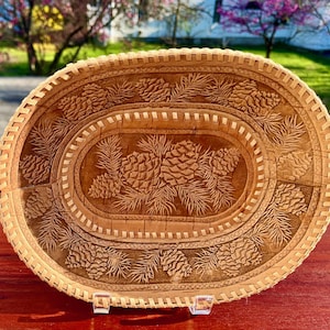 May include: Oval-shaped, hand-carved birch bark tray with a natural finish. The tray features a detailed relief of pine cones and needles, with a decorative border. The tray is supported by clear acrylic feet.