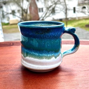 May include: A ceramic mug featuring a multi-toned glaze that transitions from a deep royal blue at the rim to a vibrant teal in the center, ending in a creamy white base. The mug has a sturdy, curved handle and a slightly textured, rounded body.