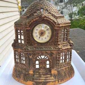 May include: A bronze-toned metal clock shaped like a building with a domed roof, featuring a circular clock face with black Arabic numerals and hands. The structure includes arched windows, a central doorway, and intricate relief detailing throughout.