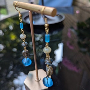 May include: Gold dangle earrings with blue, brown, and white beads. The earrings are displayed on a gold stand.