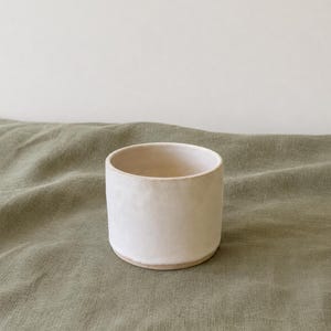 Op de afbeelding: Een kleine, cilindervormige keramische beker met een matte, gebroken witte afwerking. Het vat heeft een gladde, licht onregelmatige textuur aan de buitenzijde en een eenvoudig, open ontwerp aan de bovenkant, wat een strakke en functionele vorm toont.