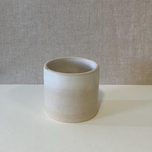 Op de afbeelding: Een cilindrische keramische plantenbak met een matte afwerking. De plantenbak heeft een kleurenschema met een verloop, dat overgaat van een lichtbeige aan de basis naar een zacht wit aan de bovenkant. De binnenkant is donkerder beige.